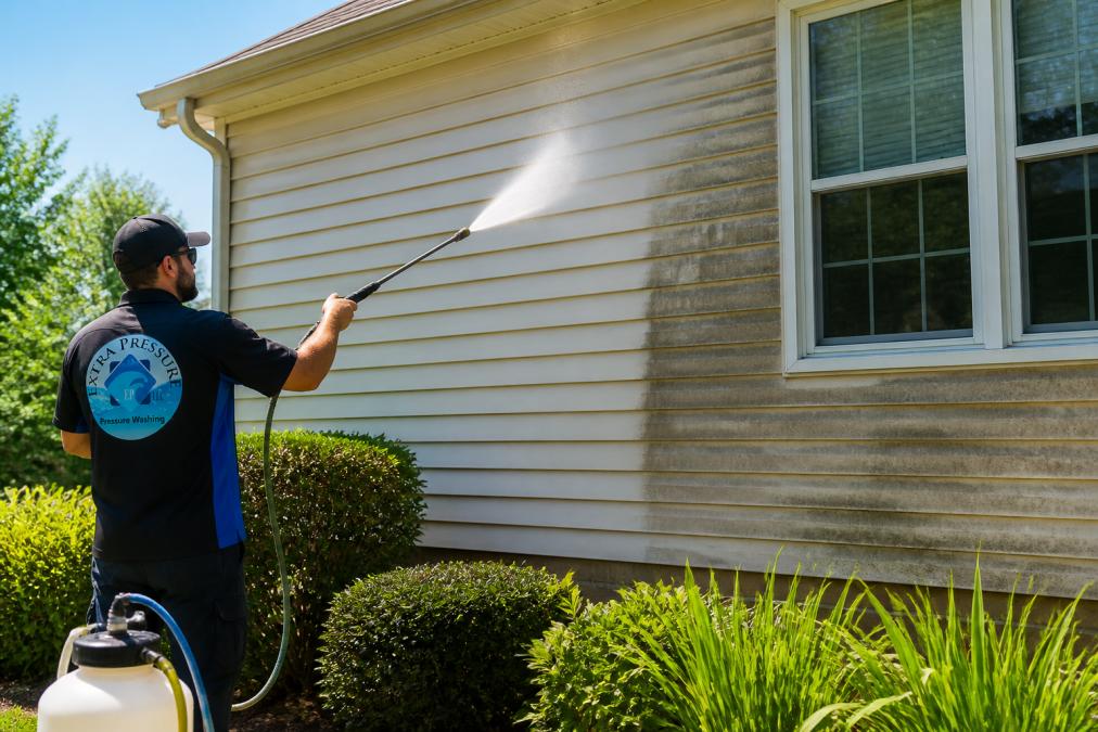 A technician in branded workwear using a low-pressure soft washing wand to clean the vinyl siding of a residential home on a sunny afternoon, with lush green landscaping visible in the foreground