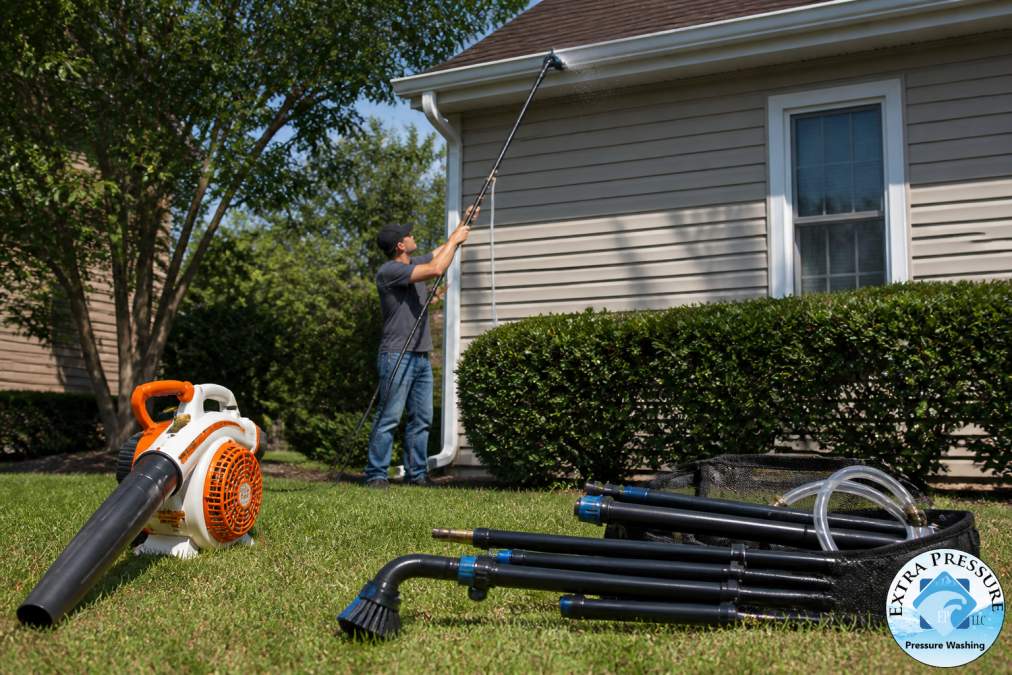 A homeowner standing on a residential lawn holding a telescopic wand attachment aimed upward at a house gutter, with a leaf blower and gutter cleaning kit laid out on the grass nearby in afternoon sunlight