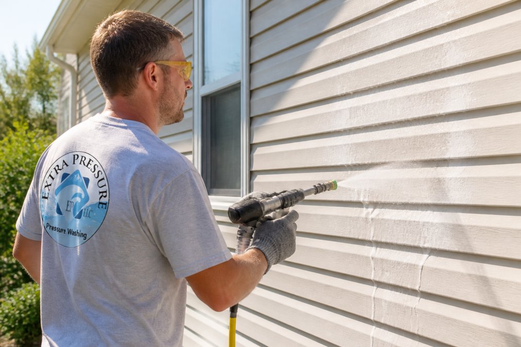 Homeowner wearing yellow safety goggles and gray work gloves applying foamy detergent solution to white vinyl siding with low-pressure spray nozzle, using vertical downward motion technique in bright morning sunlight