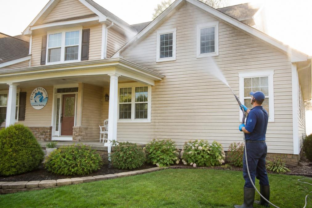 A professional exterior cleaning technician in safety gear using a soft wash wand on the siding of a two-story suburban home, with clean landscaping visible in the foreground