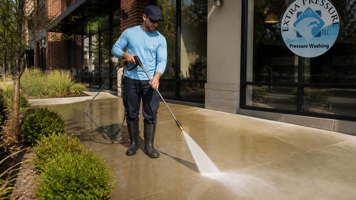 A commercial sidewalk in front of a storefront being cleaned by a professional with a pressure washer, water spray visible, bright daylight, clean pavement in the foreground